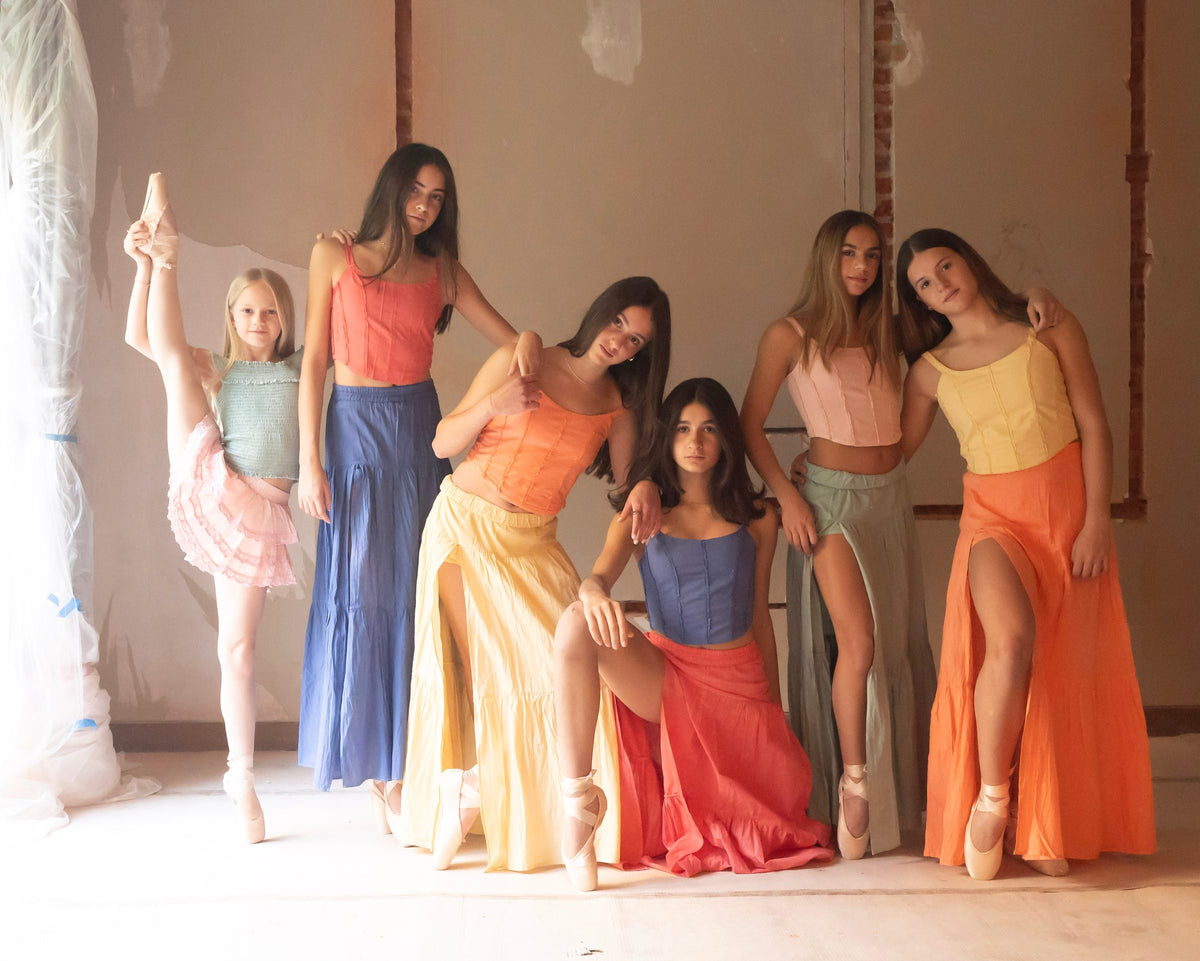 Grupo de seis con faldas largas de colores, una pierna elevada al frente — campaña Bailarinas, Amor Amore