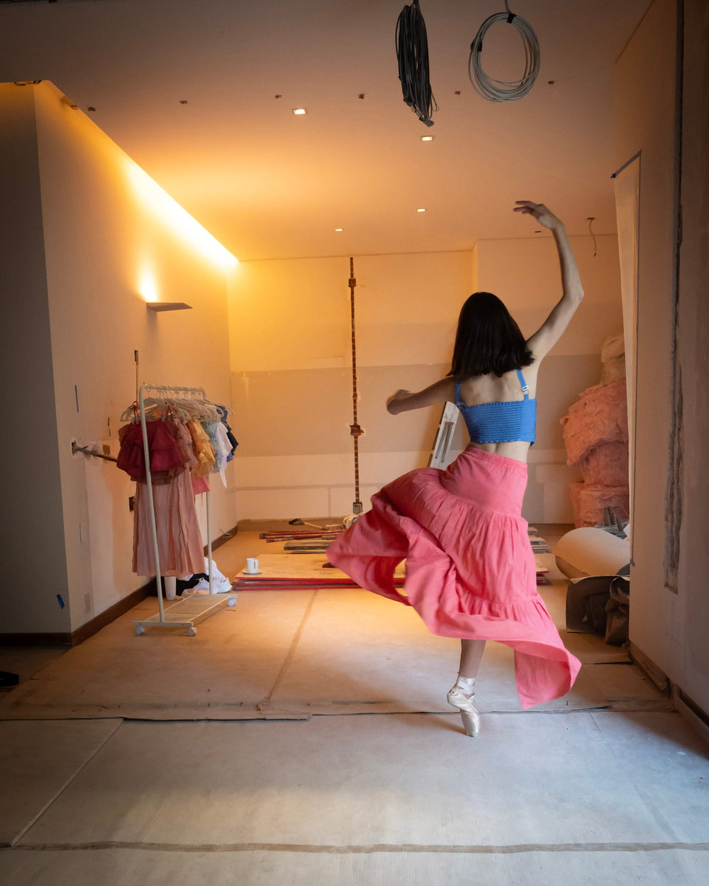 De espaldas en giro, falda larga rosa al vuelo y top azul de tirantes — campaña Bailarinas, Amor Amore