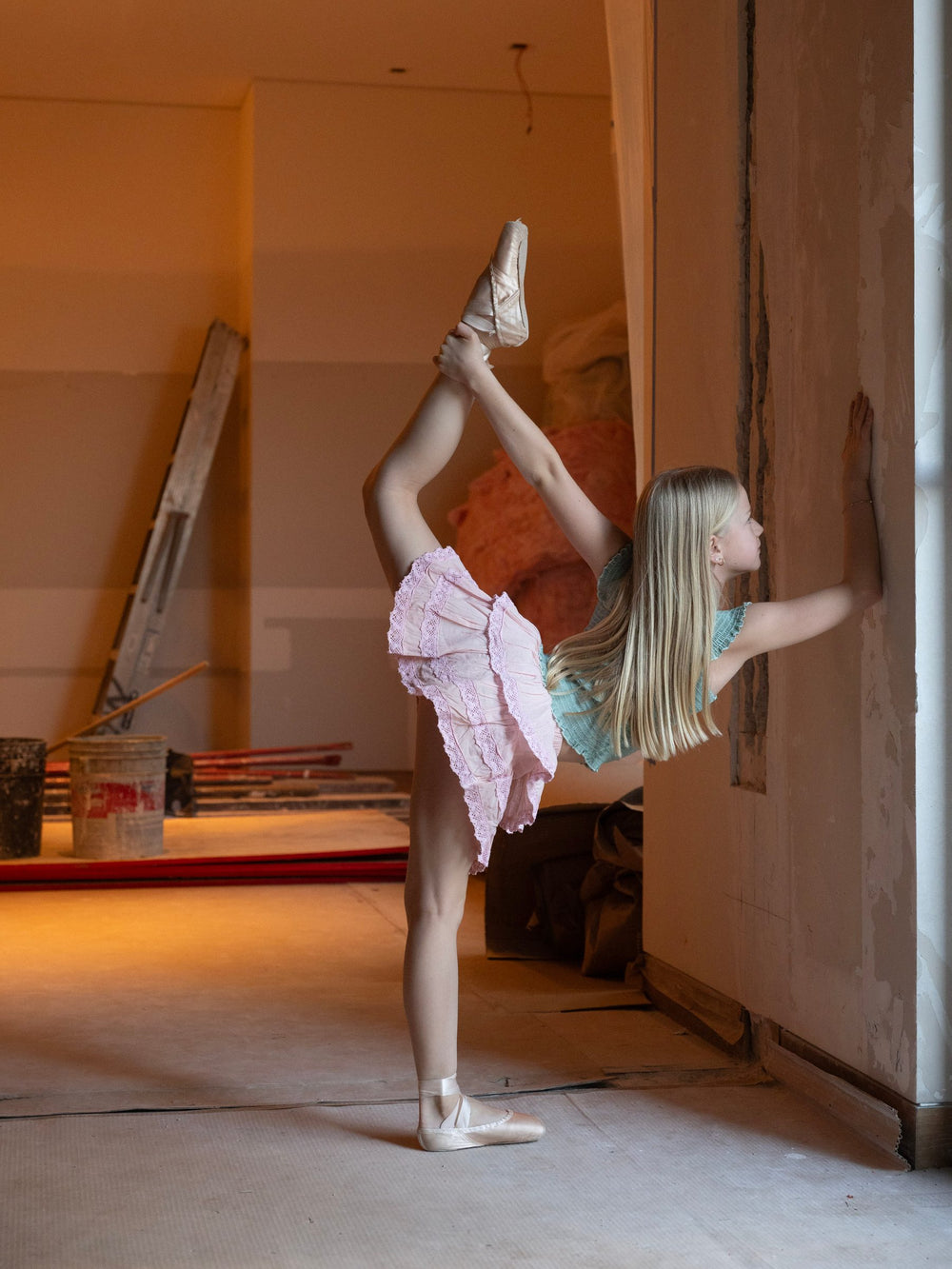 Split vertical contra el muro, falda corta de olanes rosa palo y top turquesa — campaña Bailarinas, Amor Amore