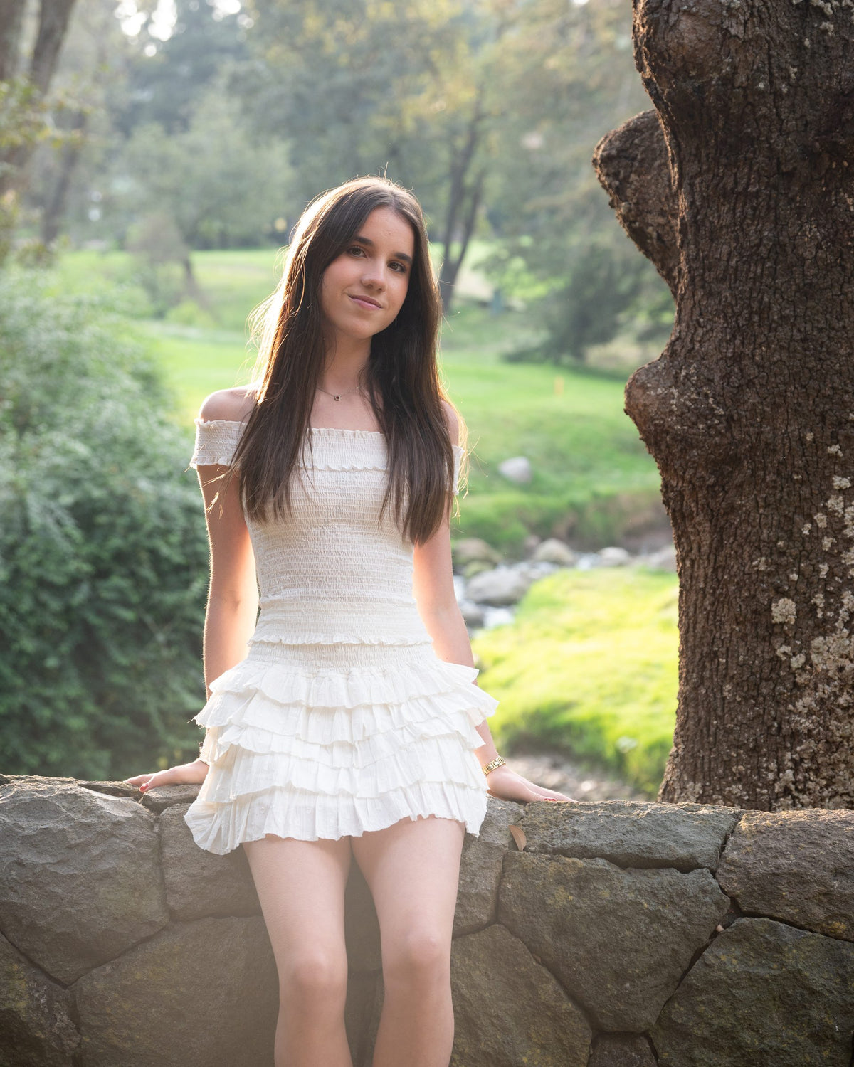 Conjunto blanco de hombros descubiertos y falda corta de olanes sobre un muro de piedra — campaña Bosque, Amor Amore