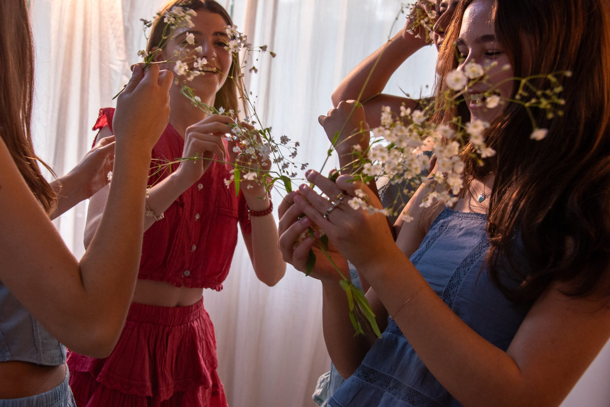 Manos con ramas de flores entre un conjunto vino y otro azul frente a la cortina — campaña Collabs, Amor Amore