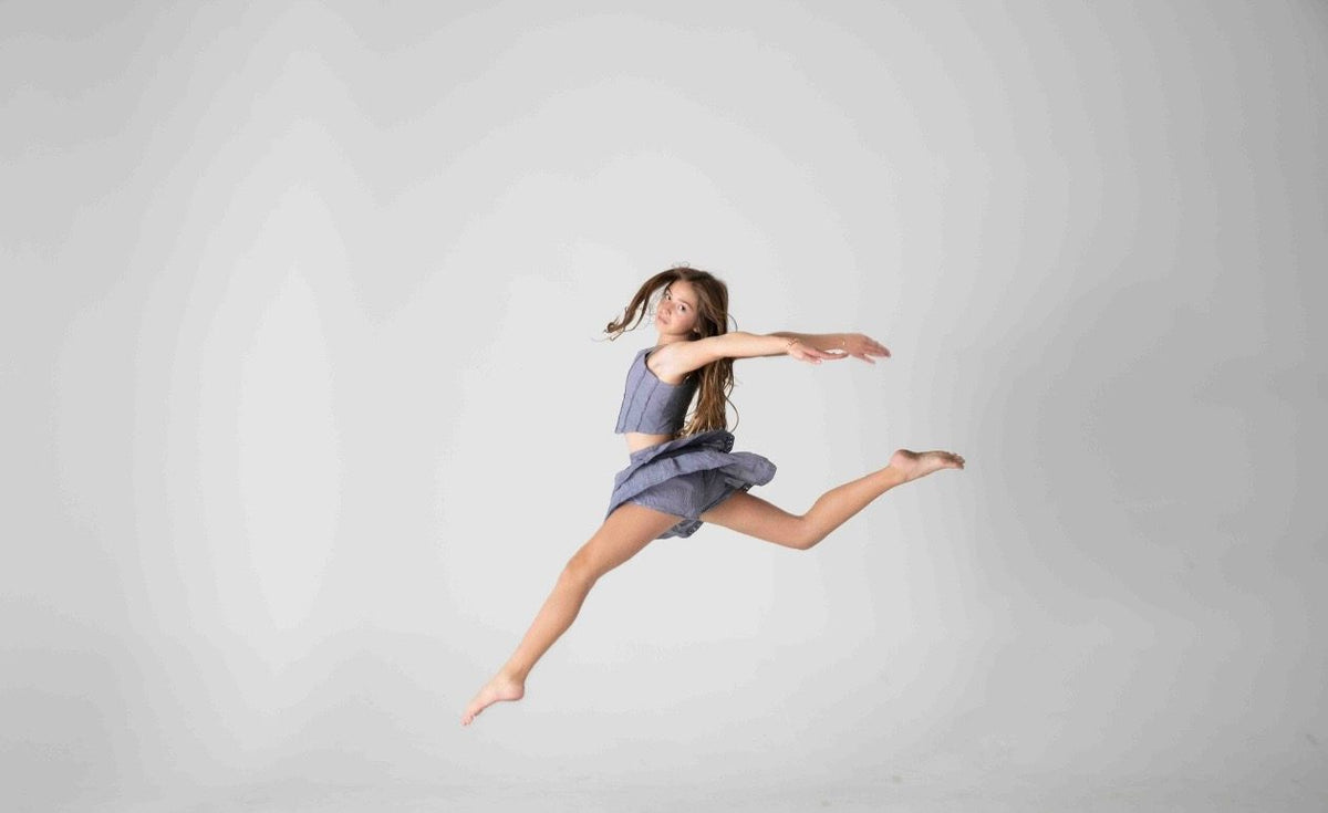 Salto de danza con conjunto azul de top de tirantes y falda corta de olanes, sobre fondo gris — campaña Estudio, Amor Amore