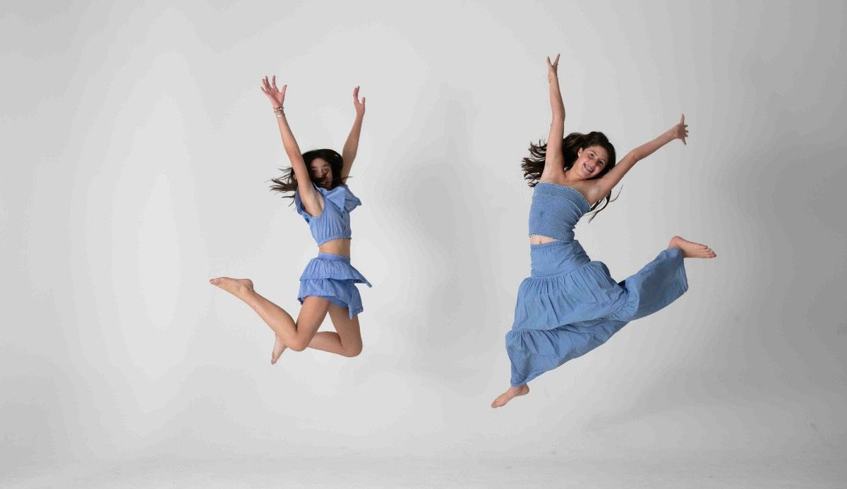 Dos saltos simultáneos en conjunto azul, falda corta de olanes y falda larga, fondo gris — campaña Estudio, Amor Amore