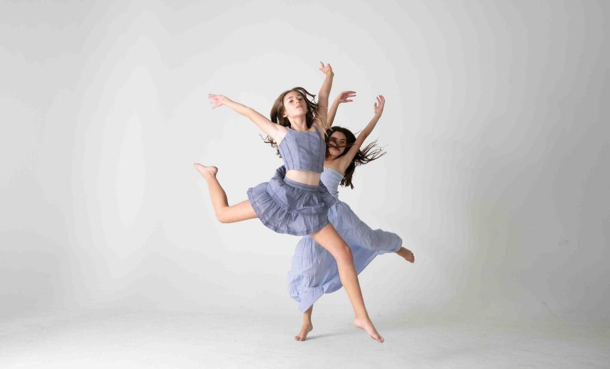 Dos figuras girando en danza con conjuntos lila de falda de olanes, fondo gris — campaña Estudio, Amor Amore