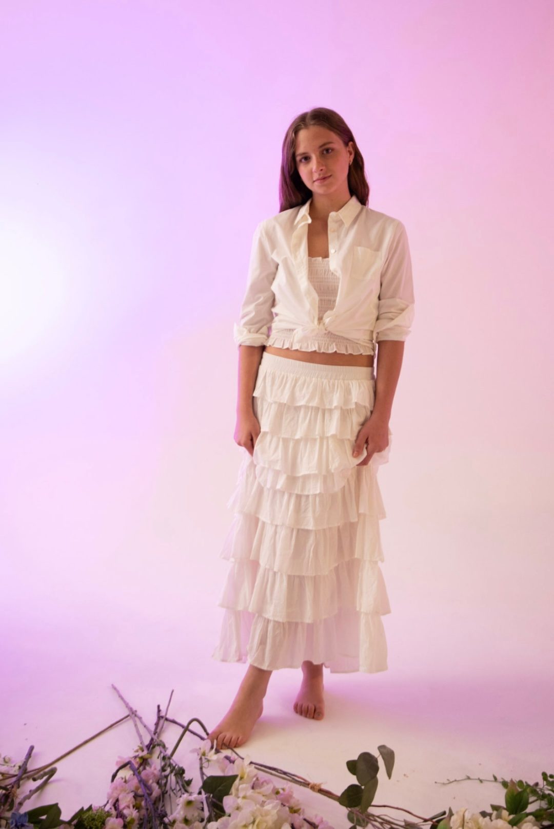 Camisa blanca anudada con falda larga escalonada blanca, de pie sobre fondo rosa palo — campaña Estudio, Amor Amore