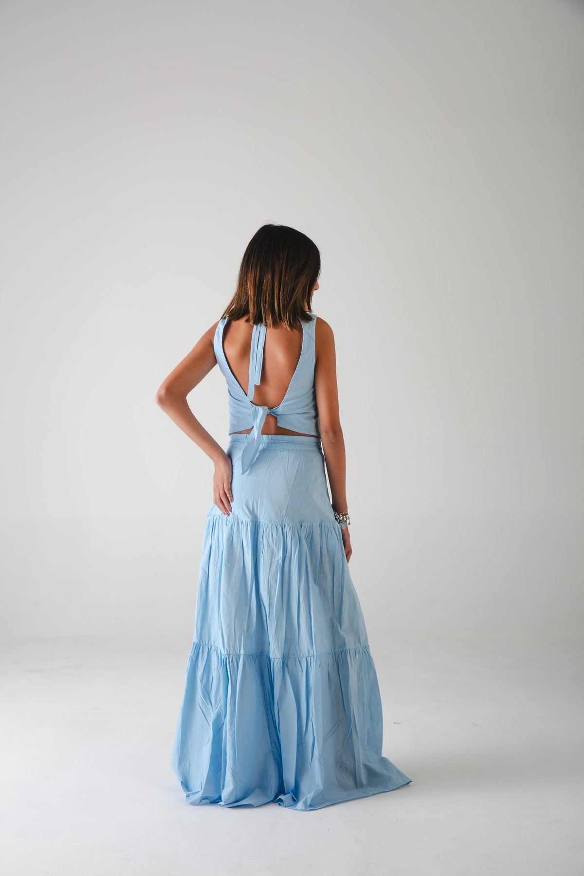 Espalda descubierta con nudo del top azul y falda larga escalonada azul, fondo gris — campaña Estudio, Amor Amore