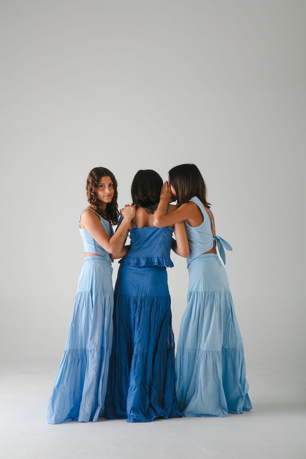 Tres faldas largas azules de espaldas, abrazadas y volteando a la cámara, fondo gris — campaña Estudio, Amor Amore