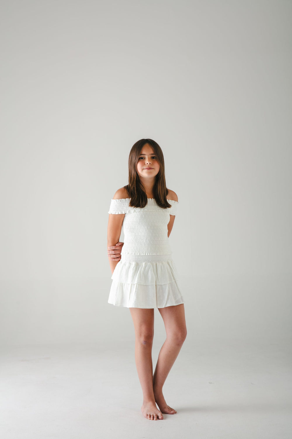 Conjunto blanco de top de hombros descubiertos y falda corta de olanes, de pie sobre fondo gris — campaña Estudio, Amor Amore