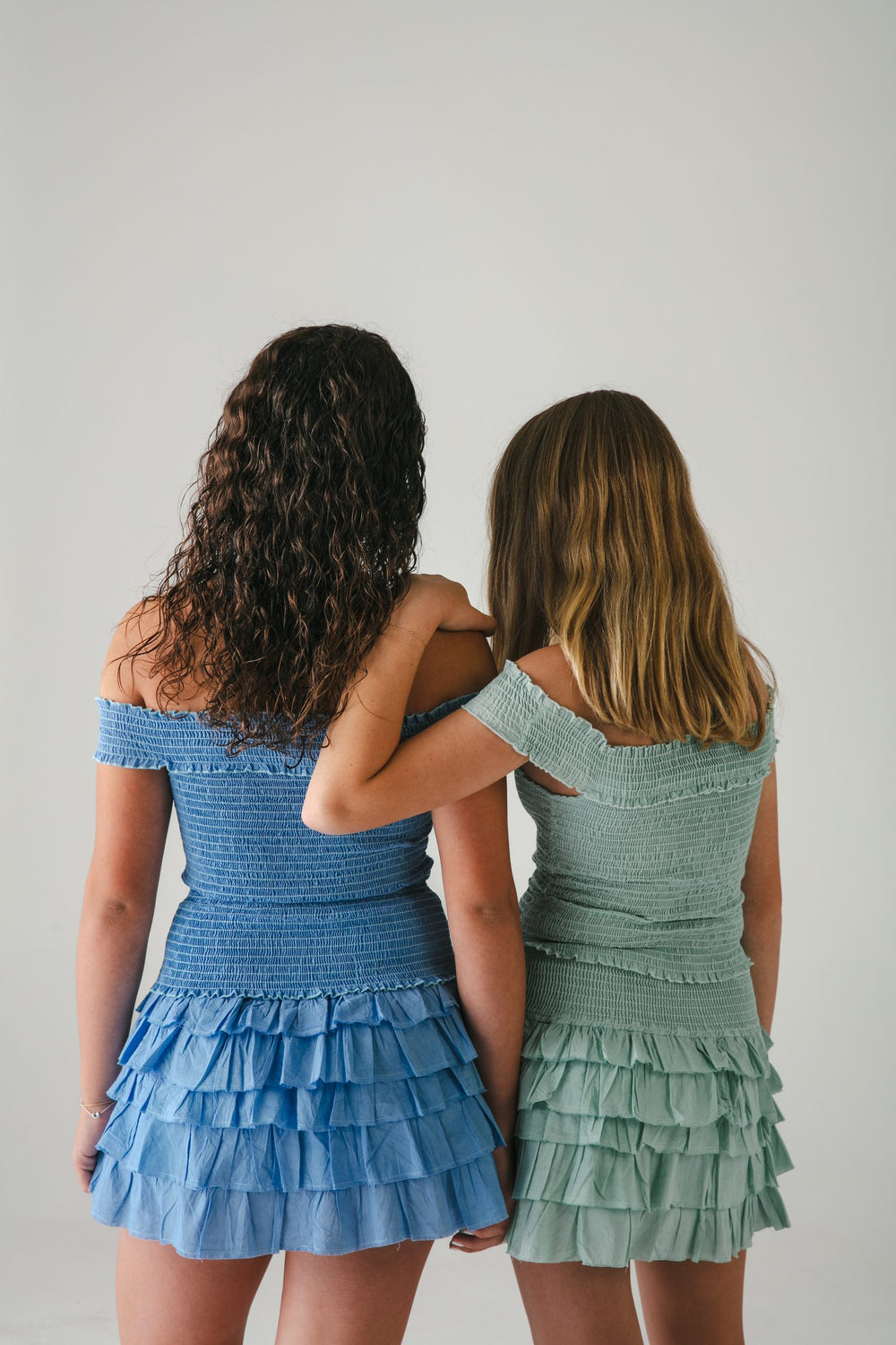 Dos conjuntos cortos azul y verde agua vistos de espaldas, tomadas del brazo, fondo gris — campaña Estudio, Amor Amore