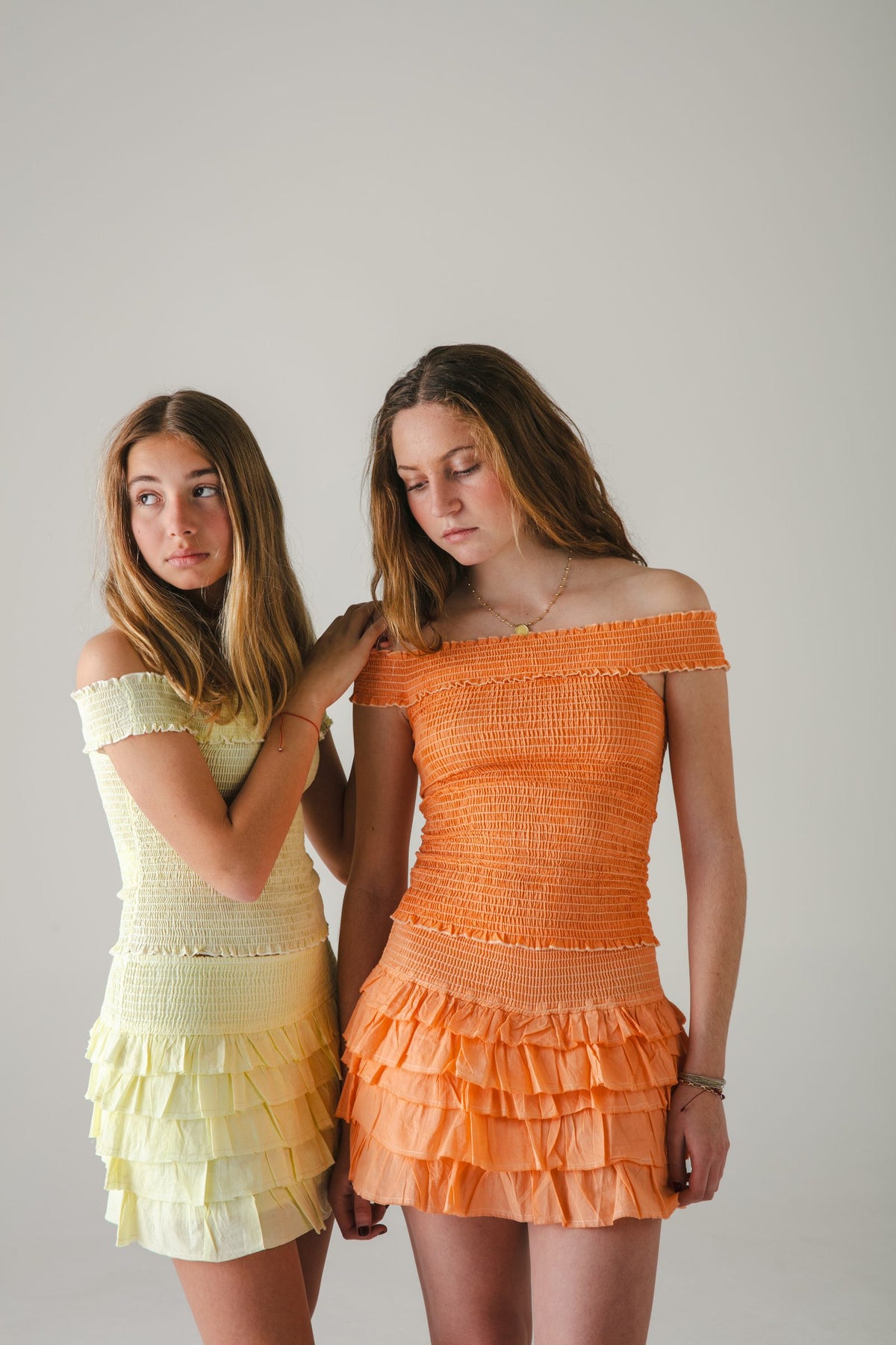 Dos conjuntos cortos de hombros descubiertos, uno amarillo y otro naranja, fondo gris — campaña Estudio, Amor Amore