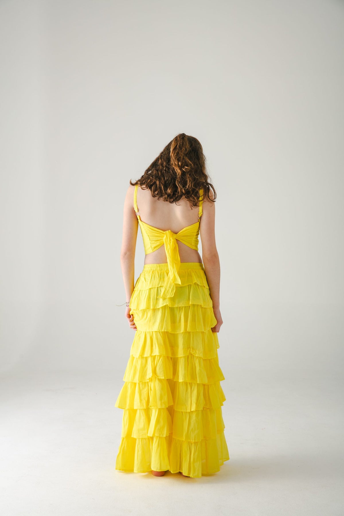 Conjunto amarillo de espaldas: top anudado a la espalda y falda larga escalonada, fondo gris — campaña Estudio, Amor Amore