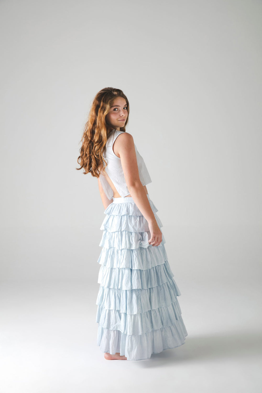 Top blanco con falda larga escalonada azul claro, girada de espaldas sobre fondo gris — campaña Estudio, Amor Amore