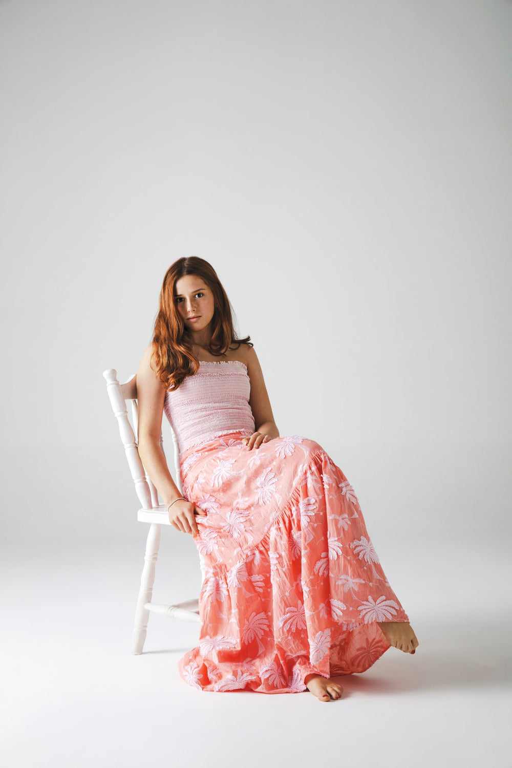 Sentada en silla blanca con top strapless rosa palo y falda larga estampada de palmeras — campaña Estudio, Amor Amore