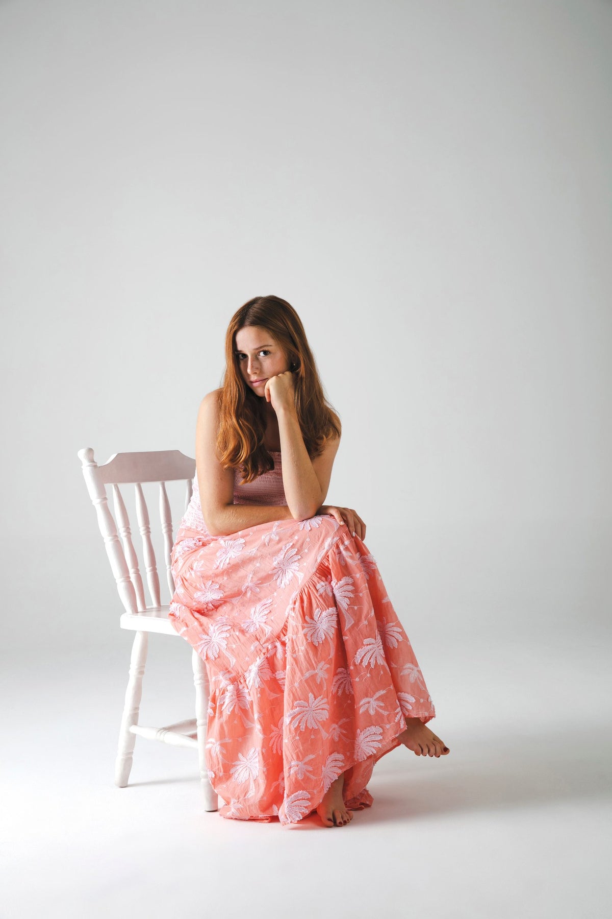 Sentada de lado en silla blanca, con la falda rosa estampada extendida sobre el piso — campaña Estudio, Amor Amore