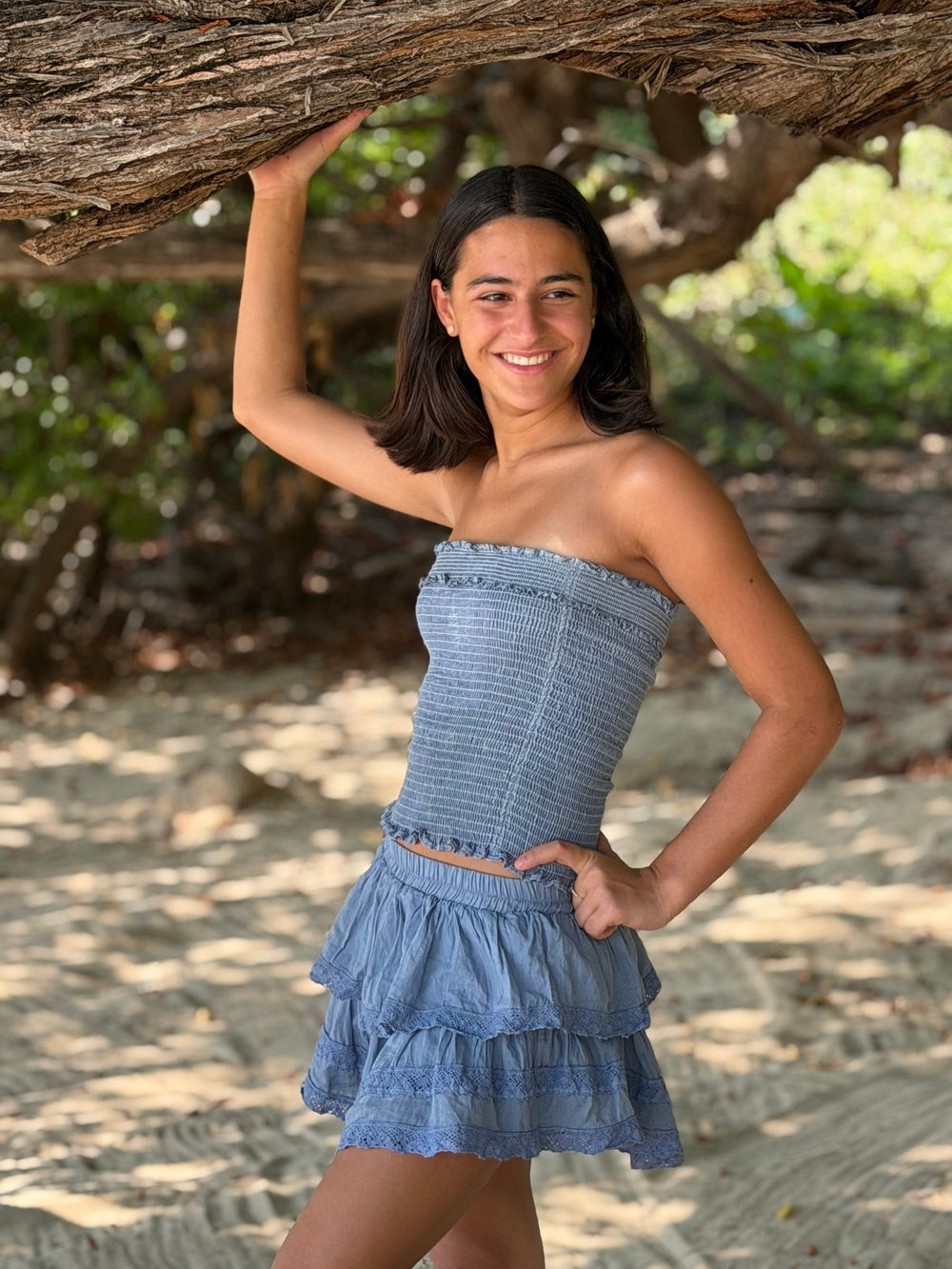 Top strapless fruncido y falda corta de dos olanes en azul, bajo la rama de un árbol en la arena — campaña Playa, Amor Amore
