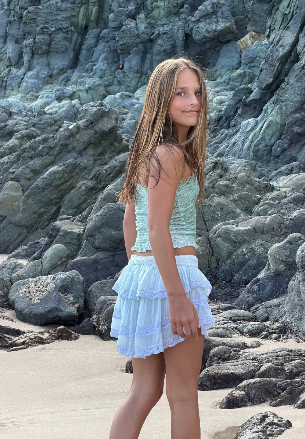 Top fruncido verde agua y falda corta blanca de olanes, de perfil junto a la roca — campaña Playa, Amor Amore