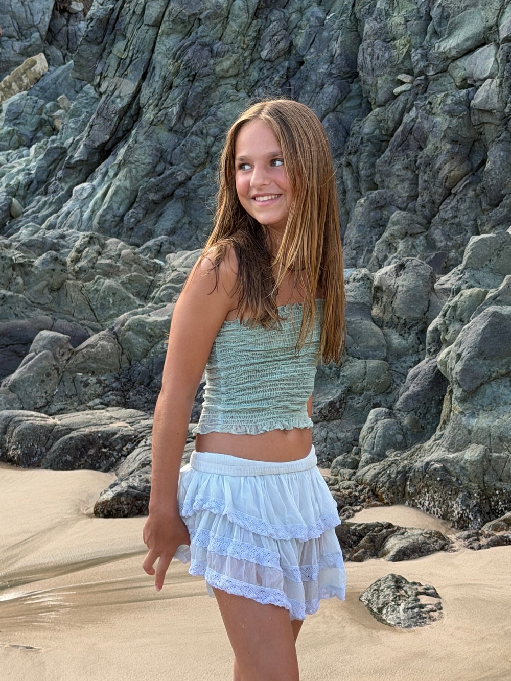 Top fruncido verde agua y falda corta de olanes con encaje, junto a la pared de roca — campaña Playa, Amor Amore